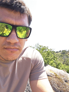 Man taking selfie in scenic outdoor setting with sunglasses, lush green trees, and bright sunny sky, capturing a moment of relaxation and outdoor adventure